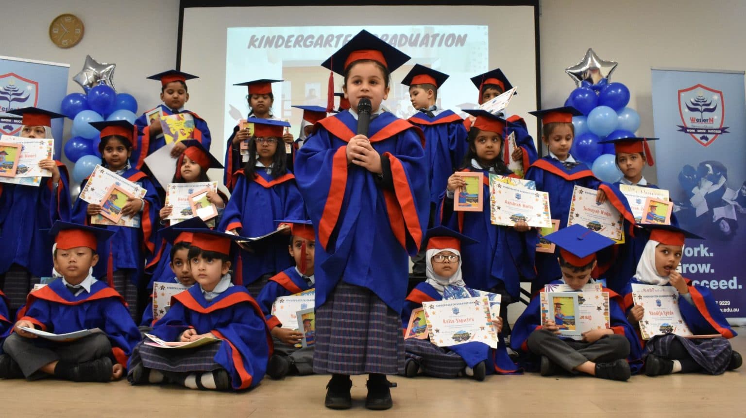 Kindergarten students in blue graduation gowns and caps holding their certificates at their graduation ceremony